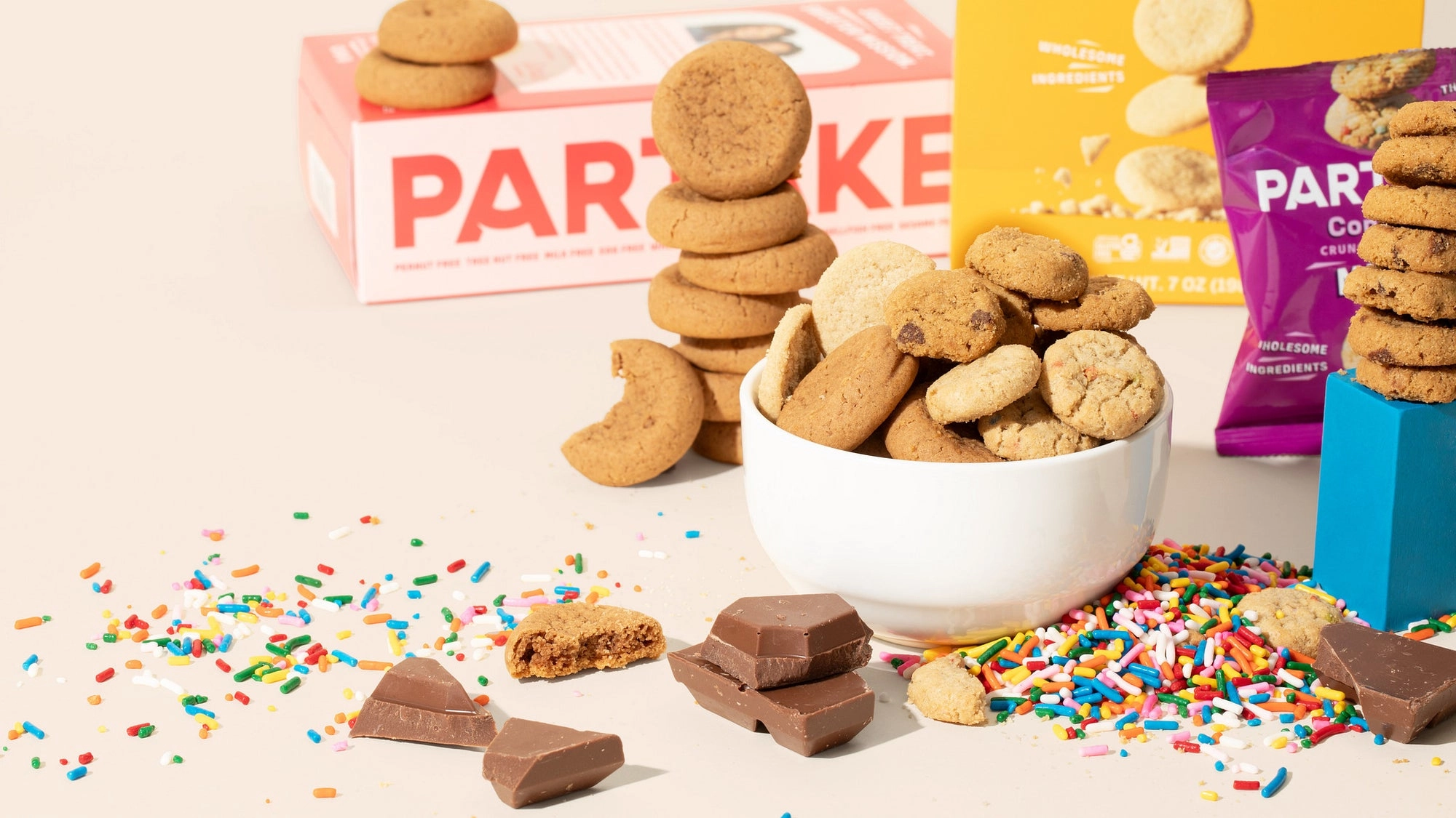 Partake cookies piled in a bowl among sprinkles and chocolate chunks, with Partake boxes behind