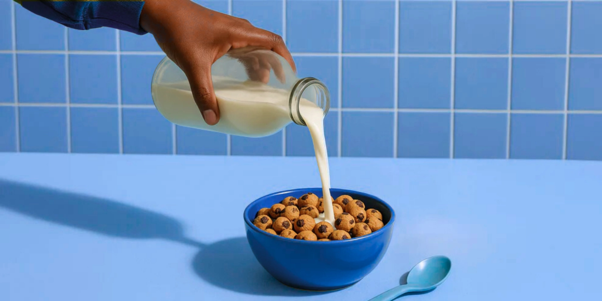 Milk poured from a glass bottle into a bowl of tiny chocolate chip cookies on a blue table